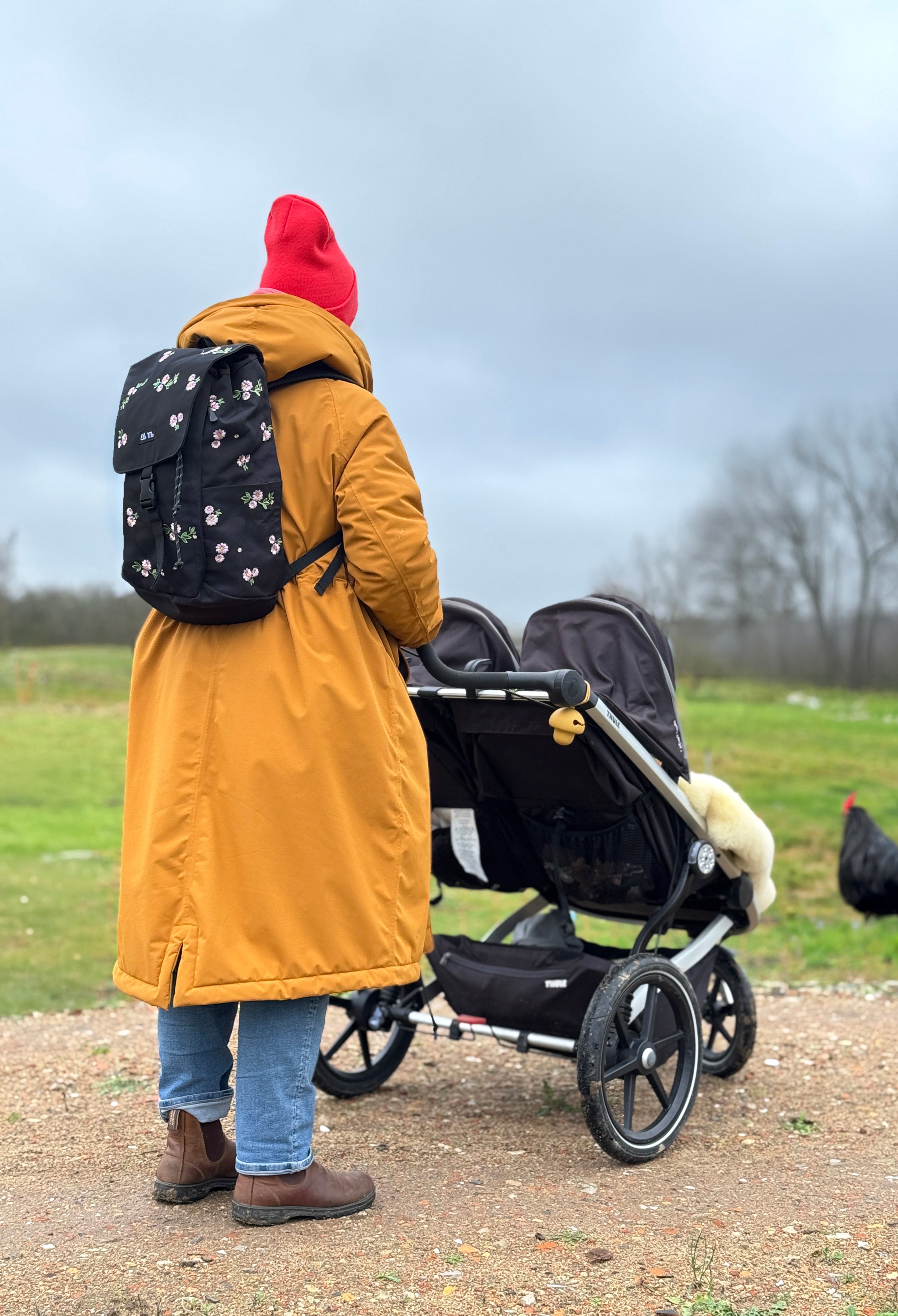 Ela Mo™ Everywhere Daypack | Embroidery Edition Upsy Daisy. Frau mit schwarzem, geblümtem Rucksack und Zwillingskinderwagen.