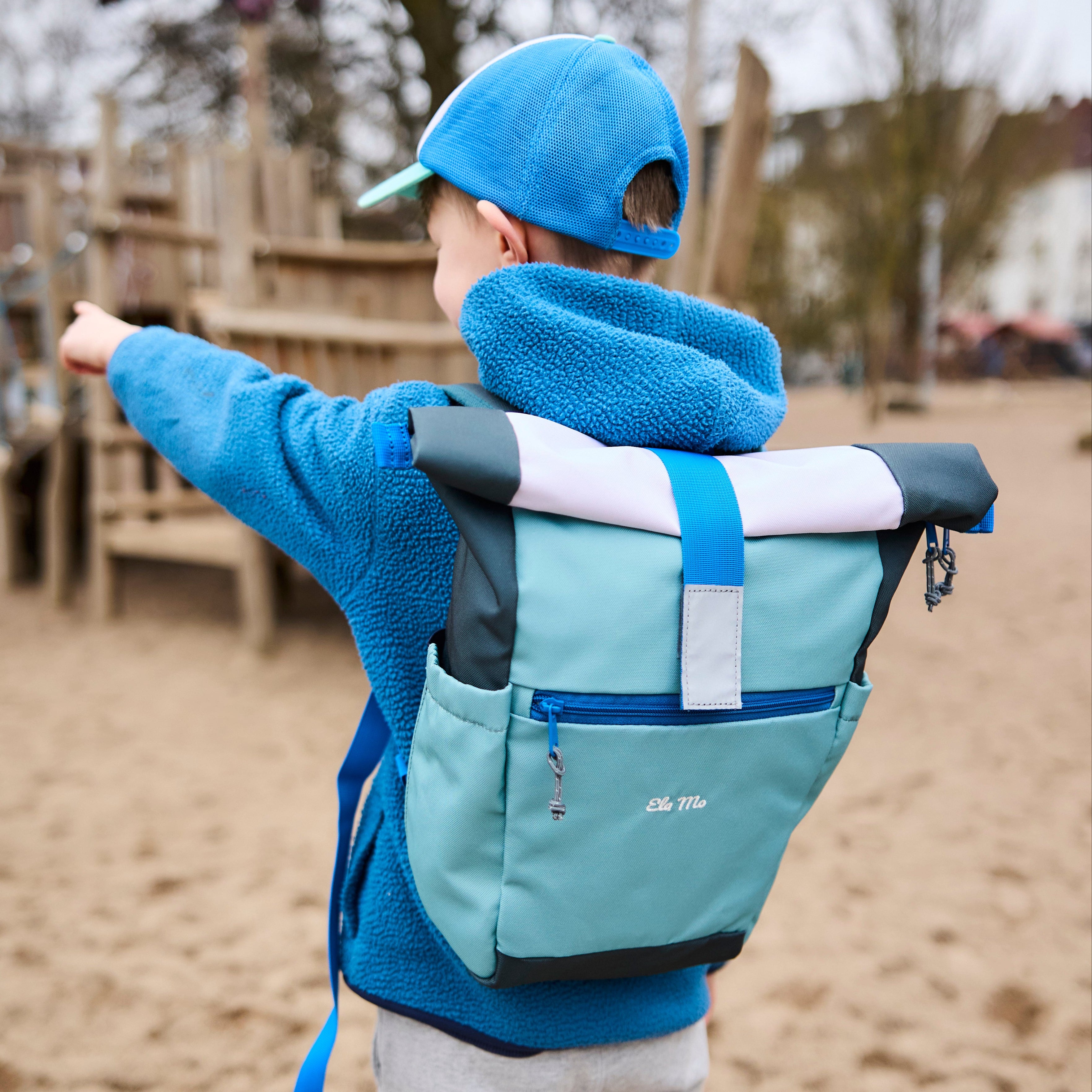 Ela Mo™ Kinderrucksack (6-10 Jahre) | Blue Mint. Ein Junge auf dem Spielplatz, der einen blauen Rucksack trägt.
