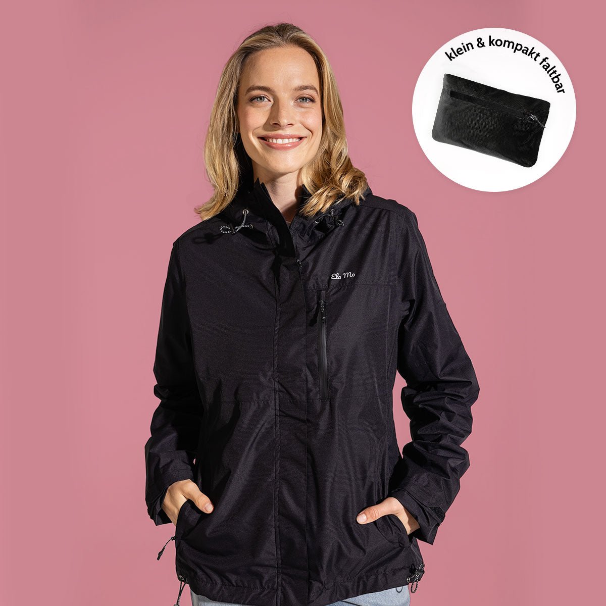 Ela Mo™ Regenjacke | Black. Eine Dame in schwarzer, kompakt faltbarer Regenjacke vor rosa Hintergrund.