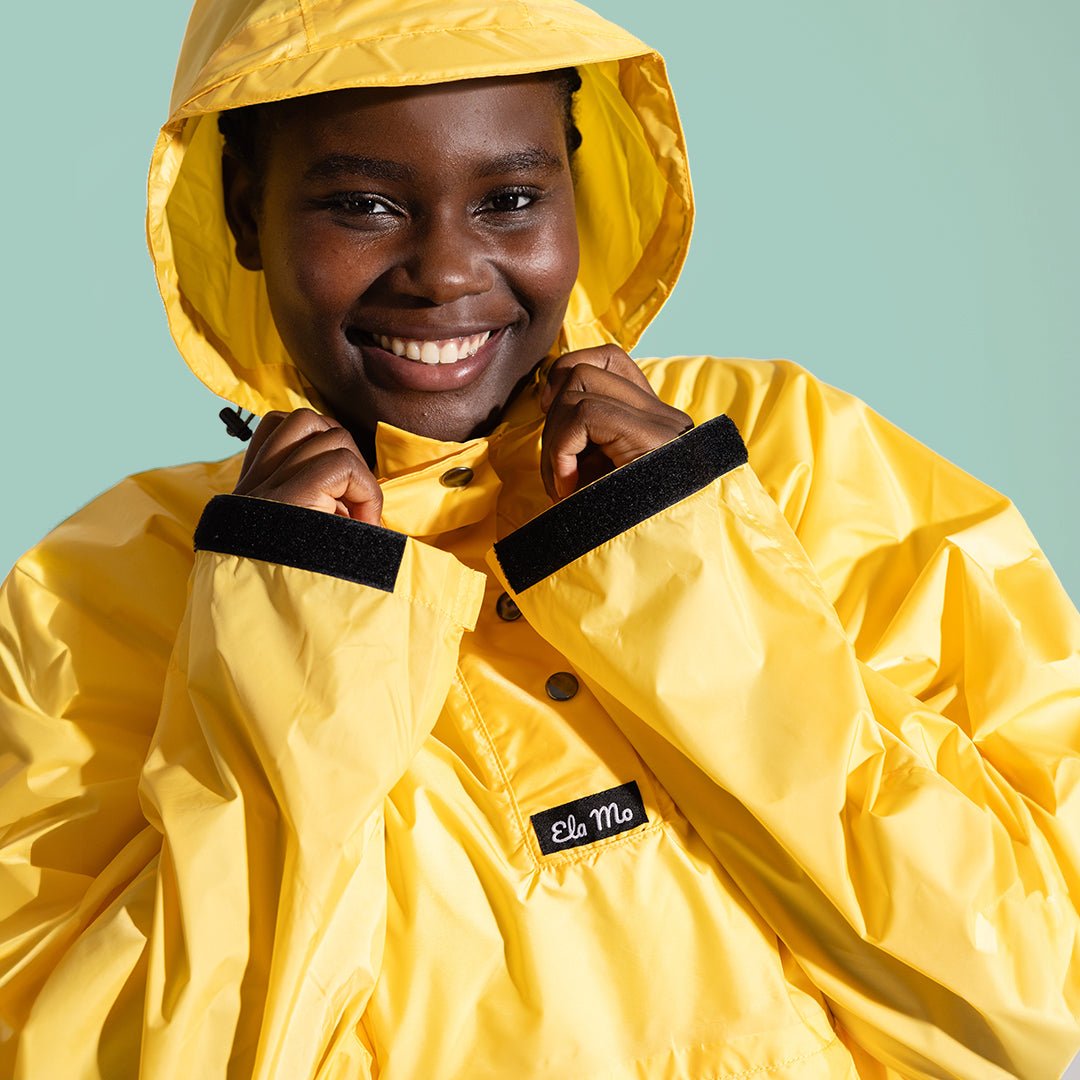 Ela Mo™ Regenponcho | Yellow. Eine Frau in einem gelben Regenponcho auf grünem Hintergrund.