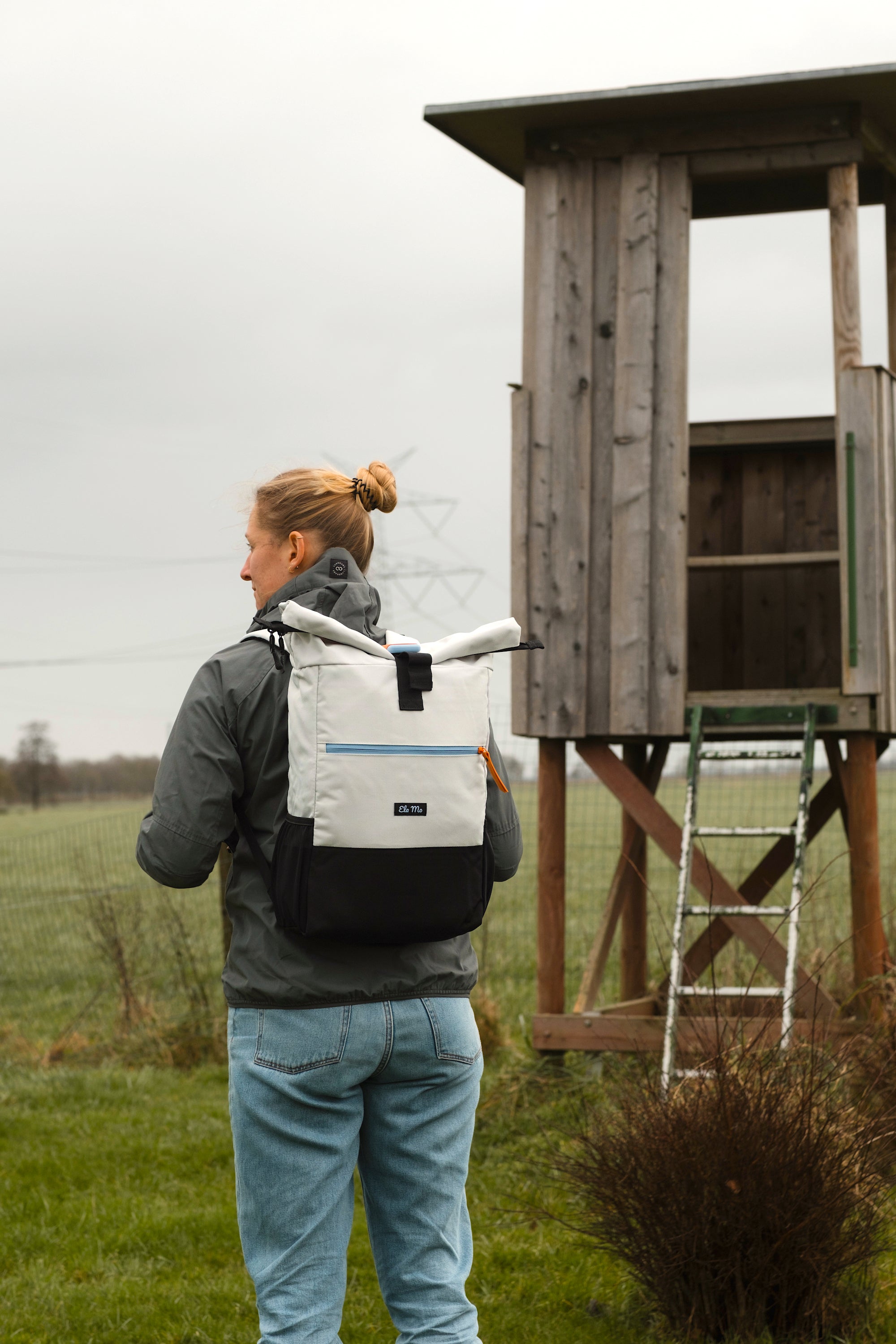 Ela Mo™ Rolltop Rucksack | Lightgrey Orange Pop. Eine Frau mit dem grauen Rucksack auf der Wiese.